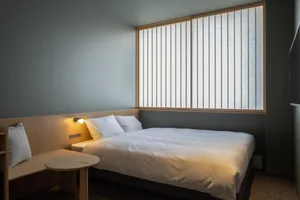 A minimalist white bedroom with a double bed, white bedding, and a small wooden table with a lamp.