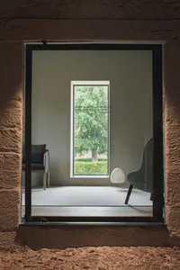Modern living room with concrete walls and floor, large window displaying greenery, minimalist furniture, and a floor lamp.