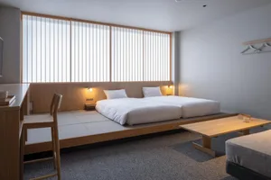 A minimalist bedroom featuring white bedding on a low wooden platform bed, a simple wooden table, and bright natural light en