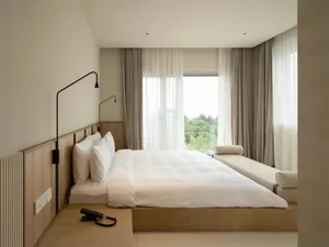 Modern minimalist bedroom with a wooden bed and white bedding, featuring a large window with sheer curtains overlooking green