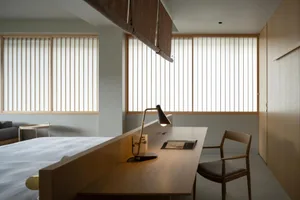 A minimalist bedroom featuring a wooden desk with a lamp, a bed, and a wooden chair.