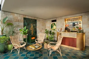 A modern living room with green and blue tiled floor, rattan chairs, and a bar area with bottles and glasses.