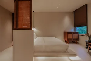 A minimalist bedroom with neutral tones, featuring a bed, side table, and cabinet with a window overlooking greenery.