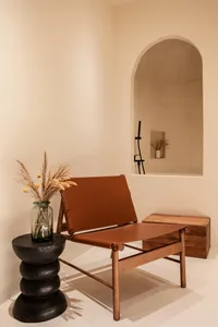 A minimalist indoor lounge area featuring a leather and wooden chair, a vase with dried plants on a black modern table, and a