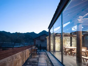 Outdoor seating area with stone walls, glass walls, and concrete patio at dusk, overlooking mountains.