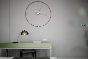 Circular modern metallic desk clock hanging above minimalist workspace with green desk and white chair.