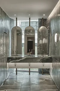 A modern public restroom with marble walls and flooring, featuring a sleek design with arch-shaped mirrors and metal fixtures