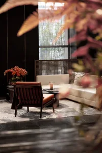 A minimalist living room setup featuring a white sofa, a wooden armchair, a coffee table with a flowerpot, and a large window
