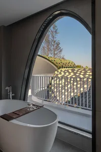 A modern bathroom featuring a white bathtub with a wooden tray and a large arched window overlooking a roof with colorful cer