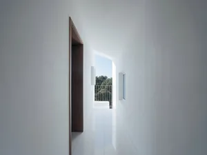 Minimalist interior design with white walls, ceiling, and floor featuring a corridor with a door and a window showing a balco