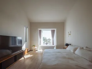 A modern bedroom with a white bed and wooden floors, featuring a large window with a view of nature.