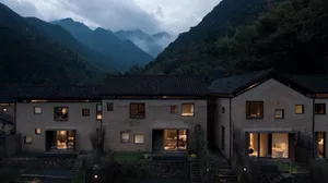 A modern house with a concrete exterior and illuminated rooms, surrounded by mountains.