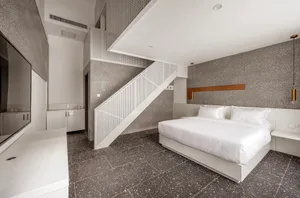 A minimalist bedroom with a white bed on a grey speckled floor, a white sink counter, and white walls with a white staircase