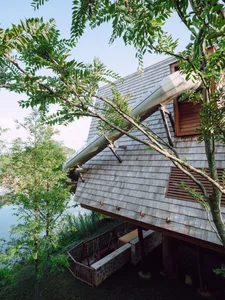 A modern treehouse with a metal slide extending from the roof, surrounded by lush greenery and a serene water body.