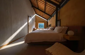 A minimalist bedroom with wooden beams, showing a bed with white bedding and a window with natural light.