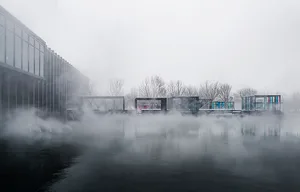 Minimalist architectural design featuring glass and concrete structures by a foggy water body.