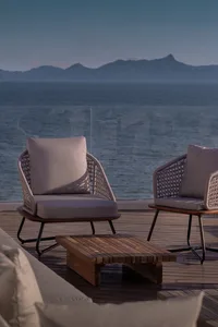 Outdoor seating arrangement featuring wooden furniture and cushions near the ocean with mountains in the background.