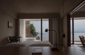 Modern minimalist bedroom with wooden furniture, fabric bedding, and metallic accents overlooking the ocean.