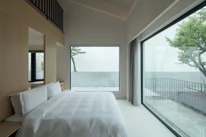 Modern minimalist bedroom featuring a glass window, white bedding, and wooden accents.