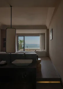 Modern bedroom with concrete walls and floor, large window overlooking the ocean, and minimalist furniture.