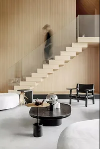 Modern, minimalist living room with wooden stairs and glass railings, featuring a round coffee table with decorative items.