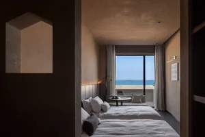 A minimalist hotel room with concrete walls and a view of the ocean through floor-to-ceiling windows.