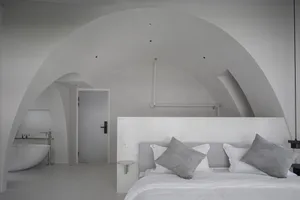 A minimalist bedroom with concrete walls, a white bed and pillows, and a sleek white bathtub in the corner.