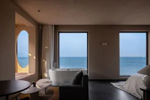 Modern hotel suite featuring a concrete bathtub, glass windows overlooking the ocean, and a cozy seating area with a sofa and