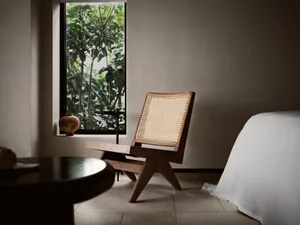 A minimalist bedroom with a wooden chair and table, natural light from a window, and a bed with a white sheet.