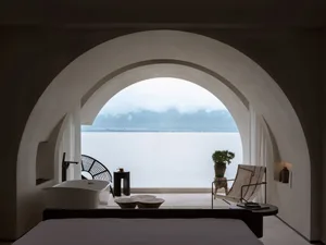 A minimalist hotel suite with a concrete floor, a bathtub, a lounge chair, and a table with a flower pot by the ocean.