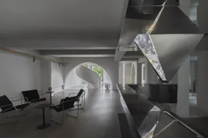 A modern lounge area with a concrete floor, metal chairs, and a glass-enclosed staircase.