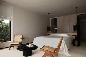 A minimalist bedroom with a large bed dressed in white linens, minimalist wooden furniture, and a black coffee table with a s