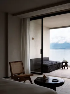 A modern, minimalist living room with a concrete floor and a large window overlooking a scenic lake view.