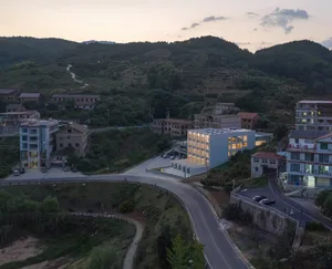 Aerial view of a modern residential building with concrete exterior and illuminated lights, surrounded by greenery and hills.