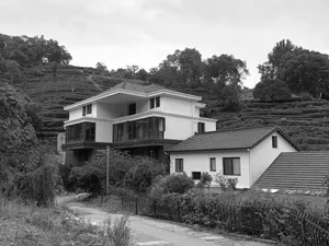 A modern residential house with a concrete exterior, surrounded by greenery and a hillside covered in terraced fields.