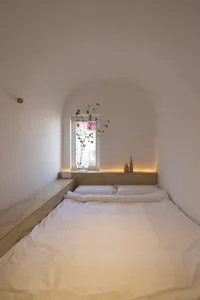 A minimalist bedroom with a white bed and wooden frame, featuring a small window with a plant and two decorative items on the