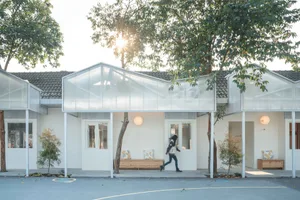A modern house with glass walls and wooden elements sits beneath tall trees.