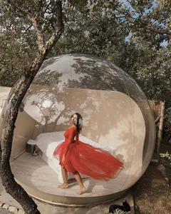 A person wearing a red dress stands inside a transparent, inflatable dome tent surrounded by trees.