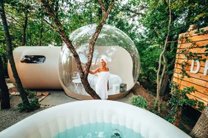 A woman wrapped in a white towel is standing next to a transparent bubble-shaped pod with a hot tub in the foreground. The po