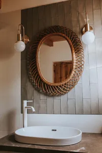 A modern bathroom with a tiled wall, oval sink, white faucet, and decorative mirror with a wicker frame.