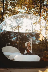 A woman sits on a transparent plastic globe bed outdoors surrounded by trees.