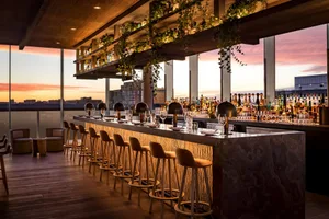 Industrial-style restaurant bar with wooden stools and glass shelves displaying bottles.