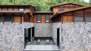 Modern residential building with wood and stone exterior, featuring large windows and a stone wall with a glass railing.