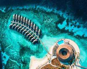 Aerial view of a resort hotel with water villas and a large circular structure in the turquoise blue water.