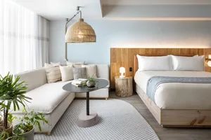 A minimalist bedroom featuring a bed with a wooden headboard, a white sofa with cushions, a side table with a plant and books