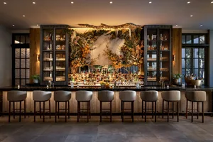 A modern bar with wooden stools, glass shelves, and plant decor.