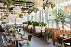 Modern restaurant interior with green plants, wooden tables, fabric seating, concrete floor and ceiling with hanging lamps.