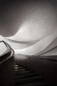 A sleek, modern staircase with curved metal steps and a dark wood railing, set against a textured, light-colored wall.