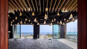 The image shows a minimalist design public space interior with wooden ceiling beams and large glass windows.