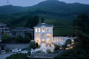 Modern residential house with white stucco and concrete exterior, set against a background of green hills and illuminated wit